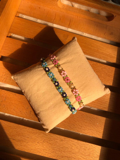 Two beaded bracelets displayed on a small tan pillow, set on a wooden slatted surface in warm sunlight.