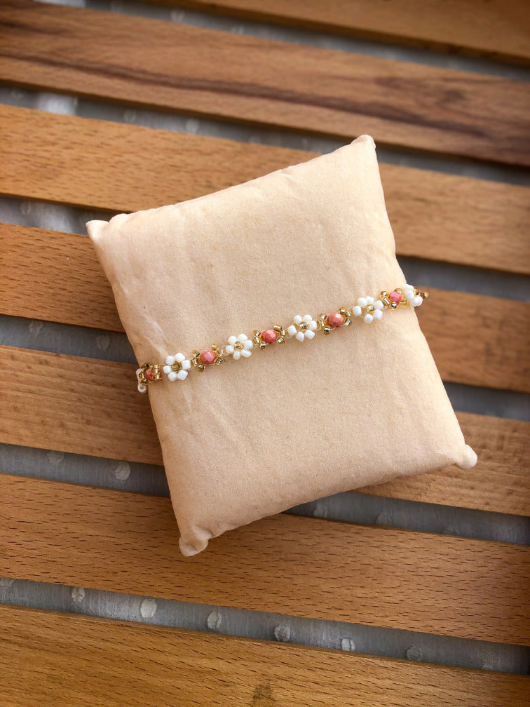 A beaded bracelet with white daisy flowers and coral-pink centers, accented with gold-toned metal, displayed on a beige fabric pillow over a wooden slatted surface.