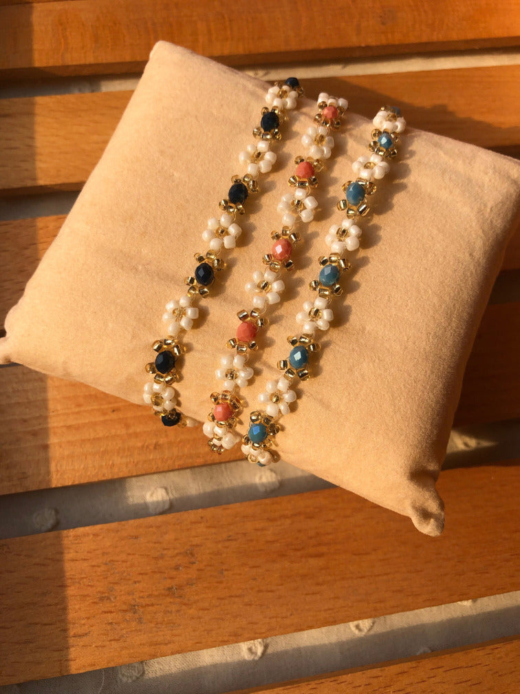 Three beaded bracelets with white beads, gold-toned beads, and colorful stone accents in blue, coral pink, and dark navy, displayed on a beige fabric pillow over a wooden slatted surface.