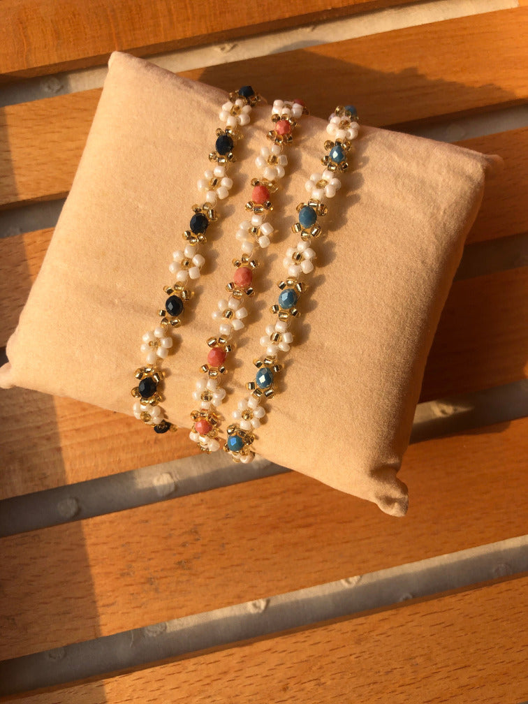 Three beaded bracelets with white beads and colorful stone accents, displayed on a beige fabric pillow over a wooden slatted surface.