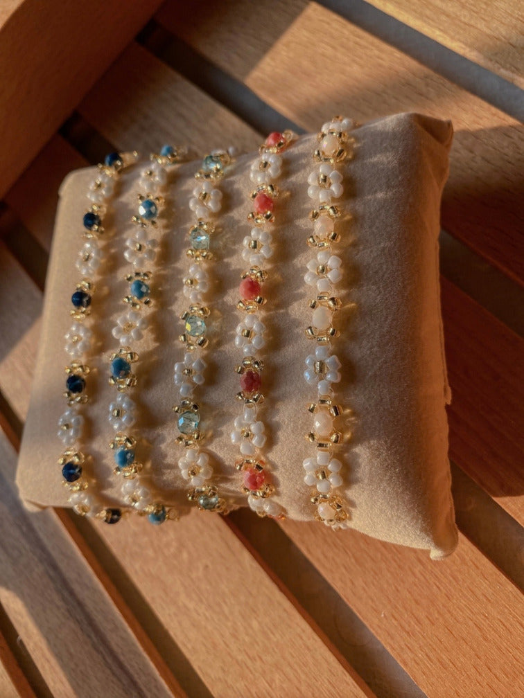 5 beaded bracelets with gold floral settings and white pearls are displayed on a beige velvet jewelry pad, set against a wooden slatted background.