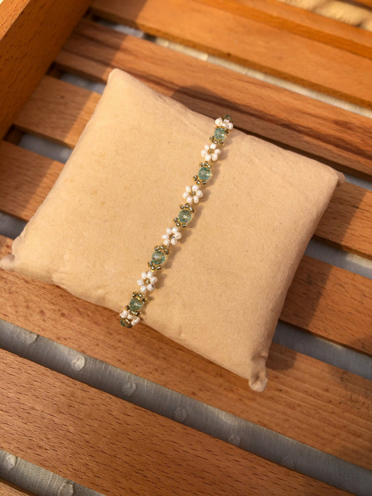 A delicate beaded bracelet with mint green and white floral motifs, displayed on a beige fabric jewelry pillow over a wooden slatted surface.