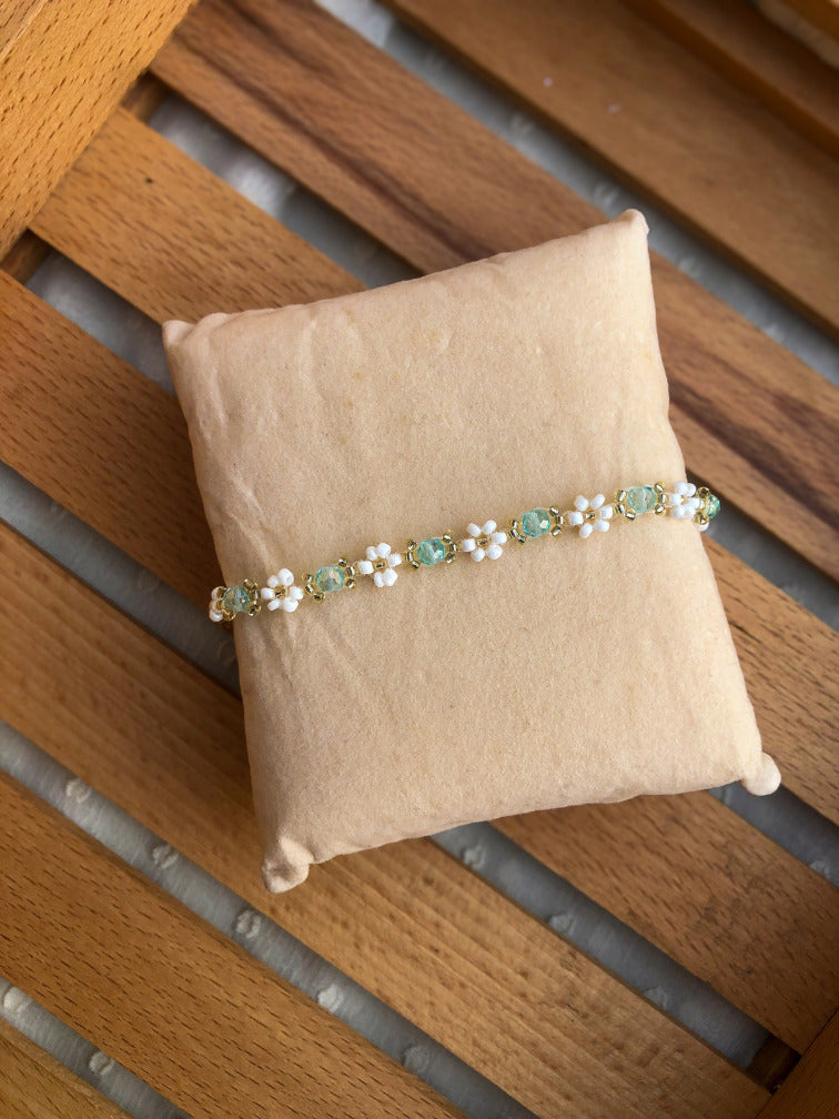 A delicate beaded bracelet with white pearl-like flowers and mint green oval beads on a gold-tone chain, displayed on a small tan fabric pillow over a wooden slatted surface.