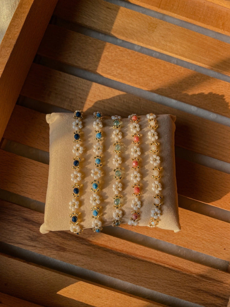 Five beaded bracelets displayed on a small cream fabric cushion, placed on a wooden slatted surface. The bracelets feature white floral beads with gold-toned settings and various colored stones in blue, green, pink, and orange.