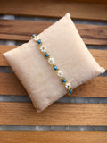 A beaded bracelet with white and sky blue beads, and gold-toned metal details, displayed on a beige fabric pillow over a wooden slatted surface.