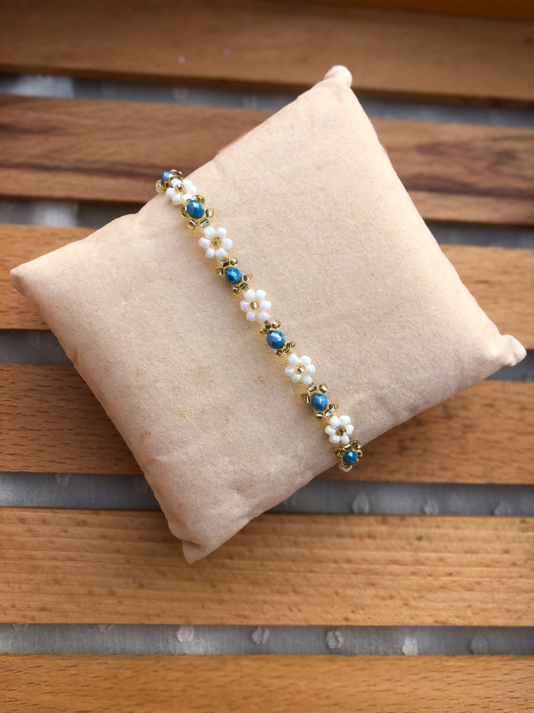 A beaded bracelet with white and sky blue beads, and gold-toned metal details, displayed on a beige fabric pillow over a wooden slatted surface.