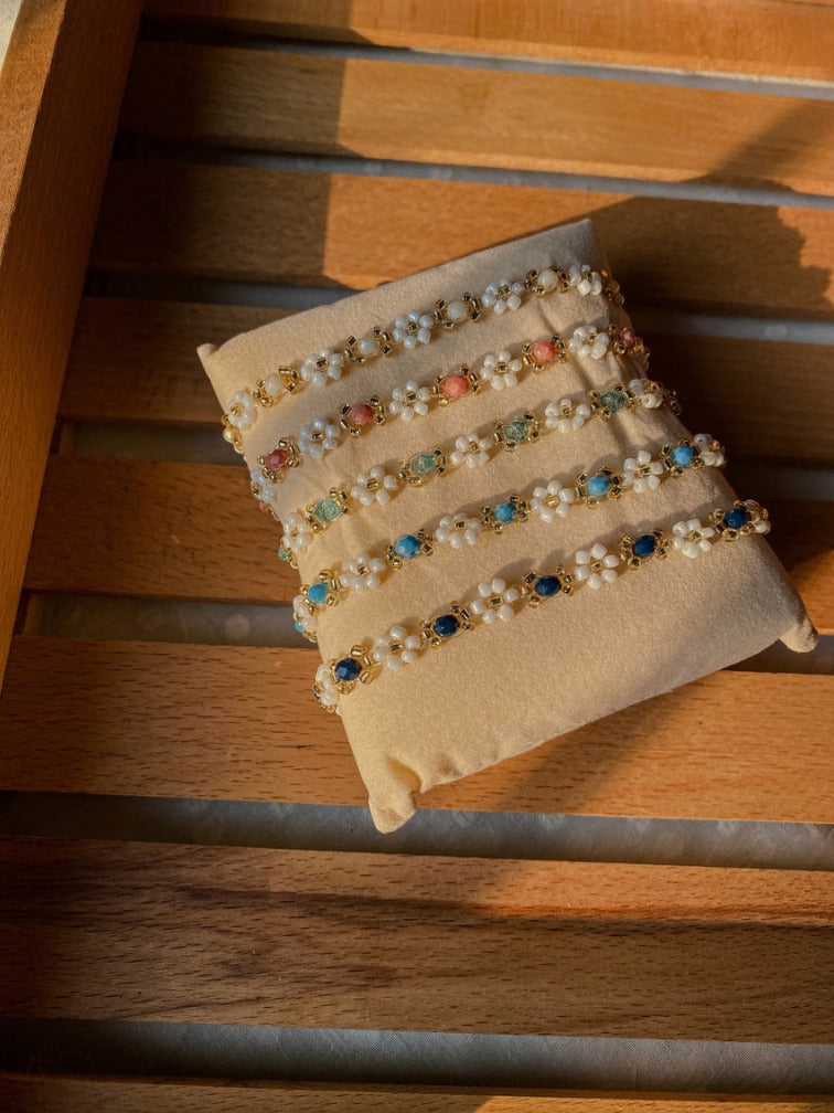 5 beaded bracelets with white pearls and colorful gemstone accents displayed on a beige fabric jewelry stand, set on a wooden slatted surface.